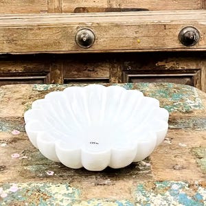 May include: A white, scalloped bowl with a flower-like design rests on a distressed wooden table. The table has a multi-coloured, weathered appearance. The background features a vintage wooden door with decorative carvings and the number 255.
