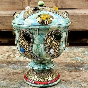 Może przedstawiać: Ozdobna, bogato zdobiona misa z pokrywką. Misa jest turkusowa i zielona ze złotymi metalowymi akcentami i kolorowymi kamieniami szlachetnymi. Podstawa jest czerwona i złota. Misa jest prezentowana na zniszczonej drewnianej powierzchni.