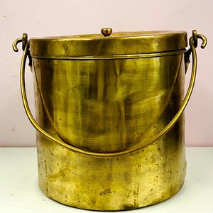 Puede incluir: Un cubo de latón vintage con tapa y asa. El cubo tiene forma cilíndrica y un aspecto envejecido y deslustrado. El asa es arqueada y la tapa tiene un pequeño pomo. El cubo está sobre una superficie blanca.