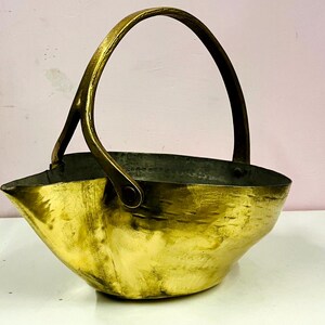 May include: A vintage brass basket with a curved handle. The basket has a rounded, asymmetrical shape with a spout-like feature on one side. The interior appears dark, contrasting with the polished gold exterior. The handle is also brass.