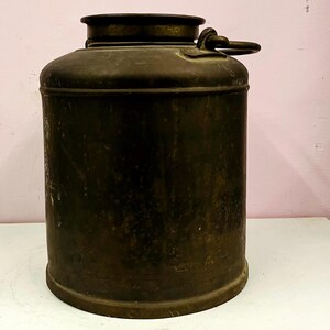 Może przedstawiać: Zabytkowy, metalowy pojemnik o cylindrycznym kształcie, z ciemną, postarzaną patyną. Posiada pokrywę z uchwytem i lekko rozszerzoną górną część. Pojemnik, prawdopodobnie wykonany z żelaza lub stali, nosi ślady użytkowania.