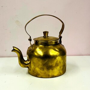 Puede incluir: Una tetera de latón vintage con un pico curvo, una tapa y un asa. La tetera tiene un acabado dorado brillante y se encuentra sobre una superficie blanca. El fondo es una pared de color rosa pálido.