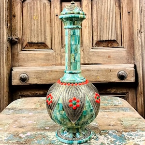 Puede incluir: Un jarrón turquesa ornamentado, con un cuerpo bulboso y un cuello largo y esbelto. Está decorado con intrincados trabajos de metal y detalles rojos y verdes. Se encuentra sobre una mesa de madera envejecida, frente a una puerta de madera vintage.