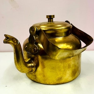May include: A vintage brass teapot with a curved spout and a hinged lid. The teapot has a handle and a small knob on top. The brass has a warm, golden color, and the teapot appears to be well-used. The background is a light pink.