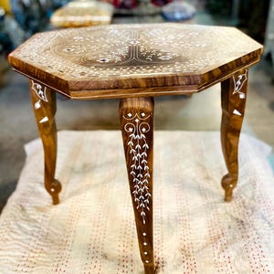 May include: A small, octagonal wooden table with detailed white inlay patterns on the surface and legs. The table has a warm, brown finish and stands on four curved legs. The tabletop features a tree-like design, with a height of approximately 40 cm.