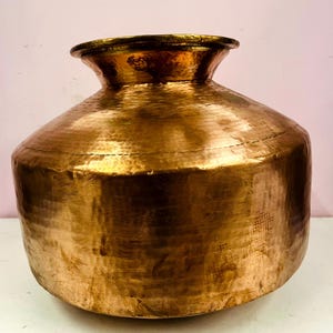 May include: A large, hammered copper pot with a wide, rounded body and a flared rim. The pot has a warm, golden-brown color and a textured surface. It sits on a white surface against a light pink background.