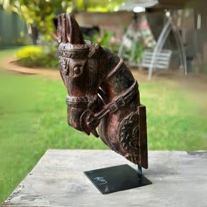 Puede incluir: Una escultura de cabeza de caballo de madera marrón oscuro con detalles intrincados. La escultura está montada en un soporte de metal negro y se exhibe sobre una superficie de madera.