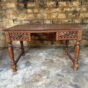 Puede incluir: Un escritorio de madera con intrincados diseños de nácar incrustados. El escritorio tiene tres cajones y cuatro patas con detalles decorativos. El escritorio es marrón con acentos blancos.