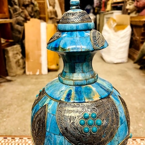 Puede incluir: Un jarrón decorativo con un diseño turquesa y latón. El jarrón presenta un cuerpo redondo y una tapa, ambos adornados con intrincados trabajos en metal e incrustaciones de turquesa. El jarrón tiene una altura aproximada de 30 cm.