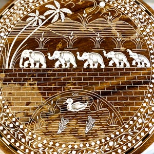 Puede incluir: Bandeja decorativa redonda de madera con un diseño intrincado. Presenta elefantes blancos caminando sobre una pared de ladrillos, con palmeras y un pato. El borde tiene un patrón floral blanco. La bandeja es marrón y blanca, con un diámetro de unos 30 cm.