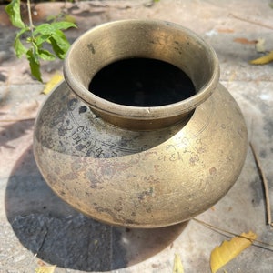 May include: A vintage brass water pot with a round body and a wide opening. The pot has a worn, aged patina and is decorated with intricate markings.