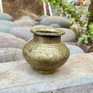 May include: A small, intricately engraved brass pot with a rounded body and a wide, flat base. The pot has a decorative design of swirling patterns and dots.