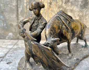 May include: Bronze sculpture depicting a matador facing a bull. The matador, in a traditional suit, holds a cape. The bull is charging forward. The sculpture is mounted on a rectangular base and has a weathered appearance.