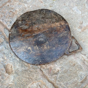 May include: A round, dark brown wooden cutting board with a small handle on the side. The board has a worn, rustic look with visible scratches and marks.
