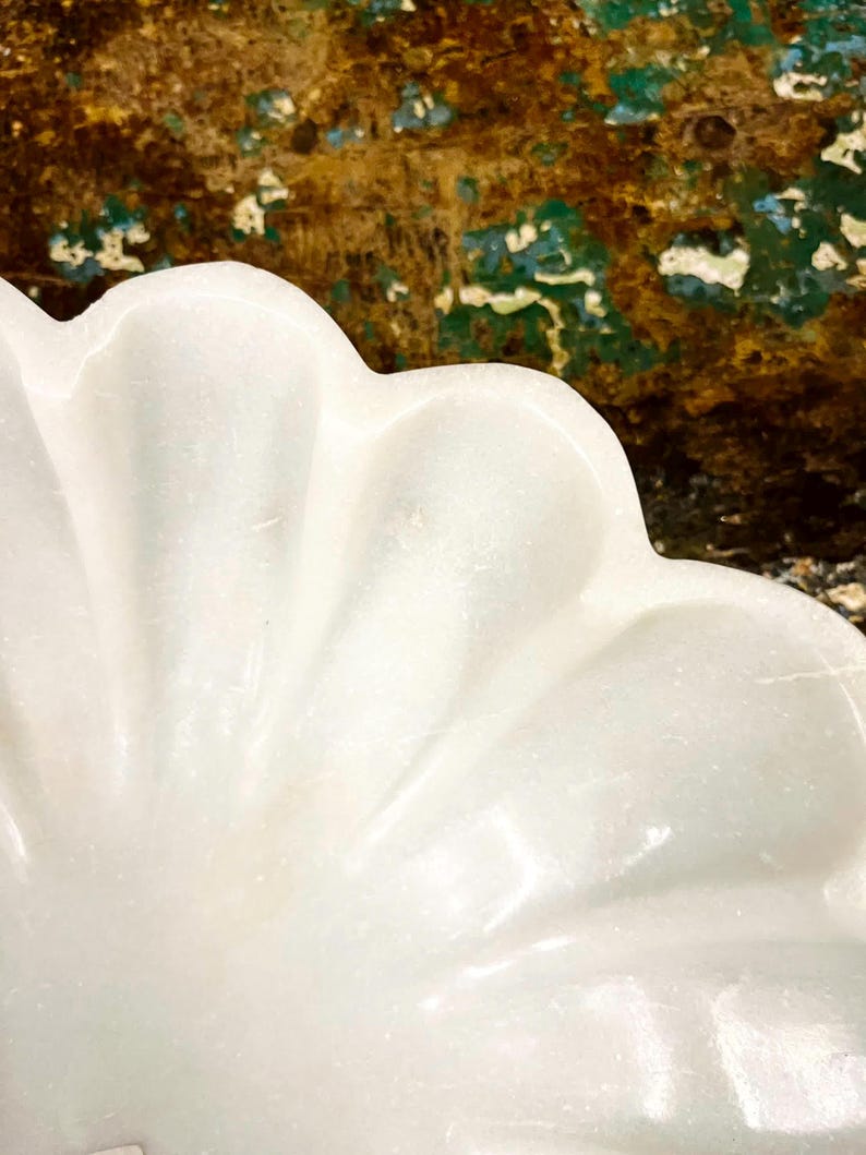 May include: Close-up of a white, scalloped-edge dish. The dish has a smooth, glossy surface and a petal-like design. The background is a textured mix of brown, green, and blue hues.
