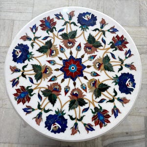 May include: A round white marble table with an inlaid floral design. The intricate pattern features blue, red, brown, and green flowers and leaves. The table's diameter is approximately 30 inches.