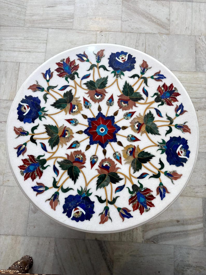 May include: A round white marble table with an inlaid floral design. The intricate pattern features blue, red, brown, and green flowers and leaves. The table's diameter is approximately 30 inches.