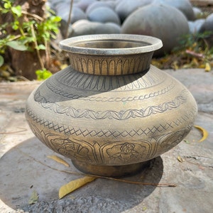 May include: A vintage brass pot with intricate etched designs. The pot features a rounded body, a flared rim, and a detailed pattern around the midsection. The brass has a warm, aged patina, suggesting its antique nature. The pot is sitting on a concrete surface.