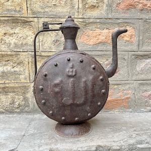 Puede incluir: Una tetera de metal vintage con cuerpo redondo y un pico largo. La tetera está decorada con un diseño en relieve y tiene una pátina marrón oscura.