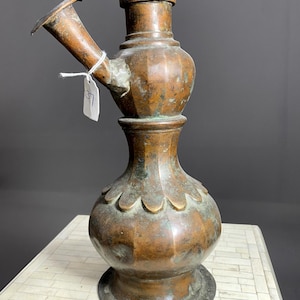 May include: A tall, copper-colored, antique hookah with a round base and a spout on the side. The hookah has a tag with the number 37 on it.