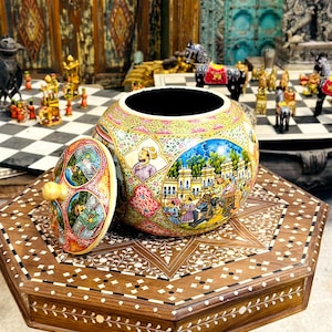 May include: A hand-painted ceramic jar with a lid, showcasing detailed artwork of figures and scenes. The jar sits on a wooden table with an inlaid design. The lid has a round knob and a painted design. The jar's measurements are in centimeters.