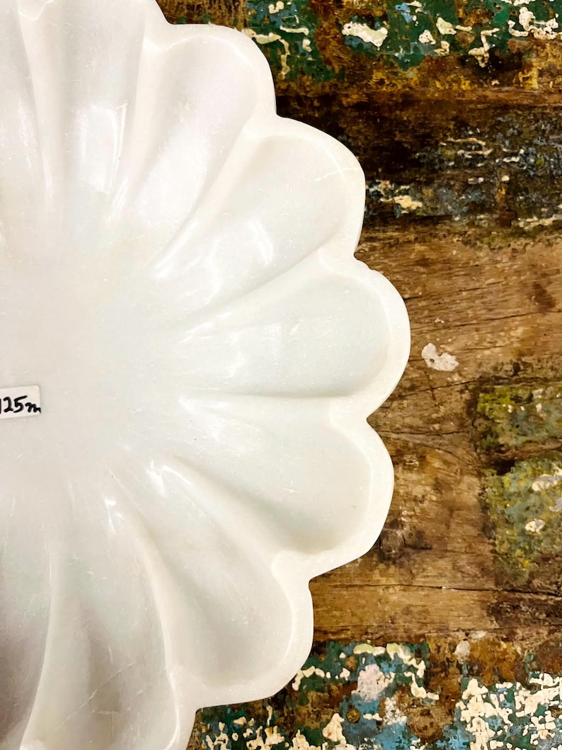 May include: A white, petal-shaped decorative dish. The dish has a glossy finish and a scalloped edge. The background is a rustic wooden surface with hints of blue and green paint.