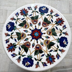 May include: A round white marble table top with an intricate floral inlay design. The design features blue, red, and brown flowers with green leaves and gold accents. The table top is a decorative piece.