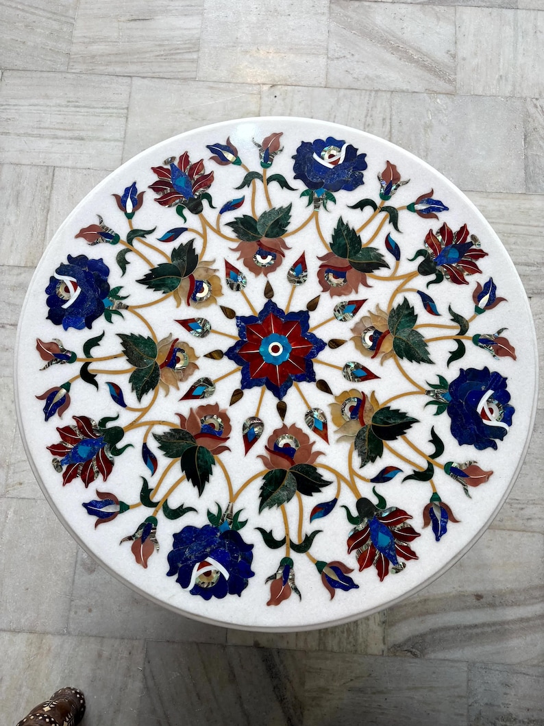 May include: A round white marble table top with an intricate floral inlay design. The design features blue, red, and brown flowers with green leaves and gold accents. The table top is a decorative piece.