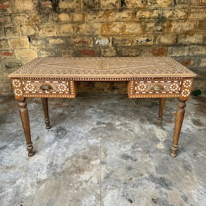 Op de afbeelding: Een rechthoekig houten bureau met ingewikkelde witte bloem- en geometrische inlegpatronen op het blad en de twee lades aan de voorzijde. Het bureau rust op vier gedraaide poten en is voorzien van twee messingkleurige metalen ladehandgrepen.
