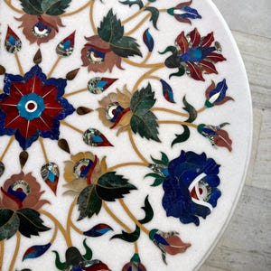 May include: A white marble table top with intricate floral inlay. The design features flowers in shades of red, blue, green, and brown, with gold-colored stems and accents. The detailed mosaic work creates a decorative surface.