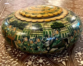 May include: A round, hand-painted decorative box with a lid. The box features a detailed scene of figures in green and white clothing, riding horses and in carriages, set against a backdrop of buildings. The lid is yellow with green floral designs.
