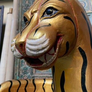 May include: A golden tiger statue with black stripes, painted with red accents around the mouth and eyes. The tiger's head is turned to the side, and its mouth is slightly open, revealing its teeth.