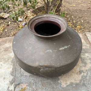 May include: A large, antique bronze-colored metal pot with a wide, rounded body and a narrow, open top. The pot has a dark interior and sits on a stone surface. The metal shows signs of age and wear.