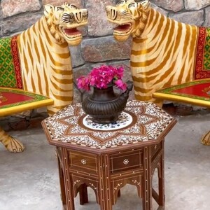 May include: Two ornate tiger-shaped chairs with green and gold patterned upholstery and red cushions, facing each other. A small, intricately carved wooden table with a mosaic top sits between the chairs. Pink flowers are in a black pot on the table.