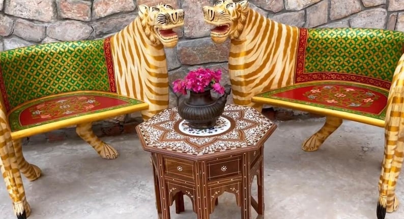 May include: Two ornate tiger-shaped chairs with green and gold patterned upholstery and red cushions, facing each other. A small, intricately carved wooden table with a mosaic top sits between the chairs. Pink flowers are in a black pot on the table.