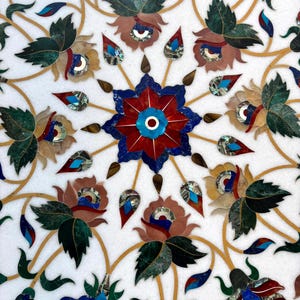 May include: Close-up of a white marble surface inlaid with a floral pattern. The design features flowers and leaves in shades of red, blue, green, and brown, with gold-colored veins. The intricate mosaic-like details create a decorative effect.