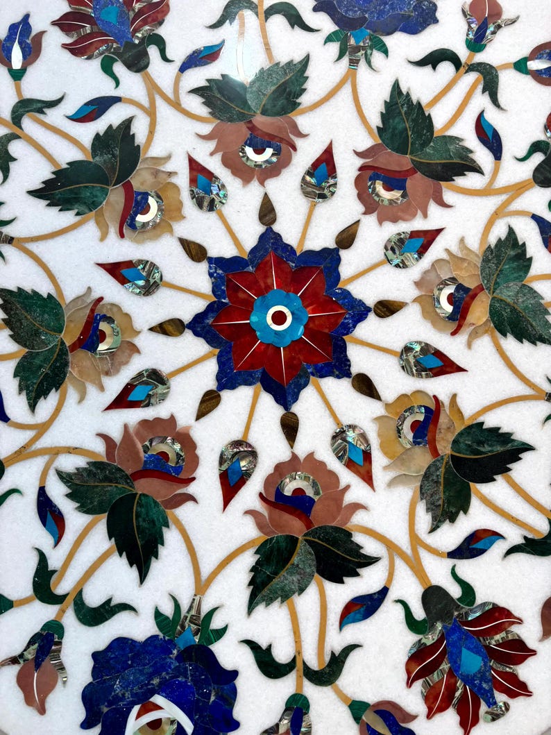 May include: Close-up of a white marble surface inlaid with a floral pattern. The design features flowers and leaves in shades of red, blue, green, and brown, with gold-colored veins. The intricate mosaic-like details create a decorative effect.