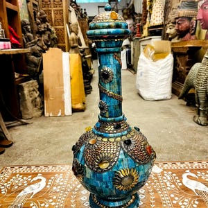 Pode incluir: Um vaso turquesa alto e ornamentado com detalhes intrincados em mosaico e metal. O vaso apresenta um corpo bulboso, um pescoço esguio e uma tampa decorativa. O design inclui padrões florais e geométricos, com detalhes dourados e de outras cores. Cerca de 60 cm de altura.