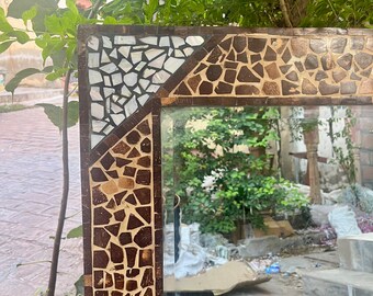 Puede incluir: Un espejo rectangular con un marco de mosaico. El marco presenta un patrón de piezas marrones, blancas y beige. El espejo refleja un fondo verde frondoso y una superficie de ladrillo.