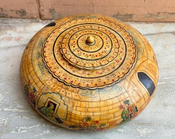 May include: A round wooden box with a painted lid. The lid features a circular design with intricate details in red, yellow, and black. The box is decorated with a scene of people and buildings in a similar style.