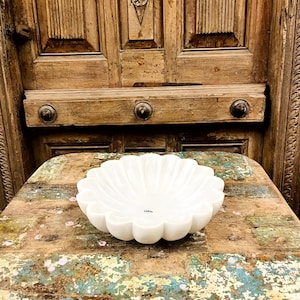 May include: A white marble bowl with a scalloped edge. The bowl has a flower-like shape with rounded edges and a smooth, polished surface. The bowl is on a rustic, multi-coloured wooden surface.