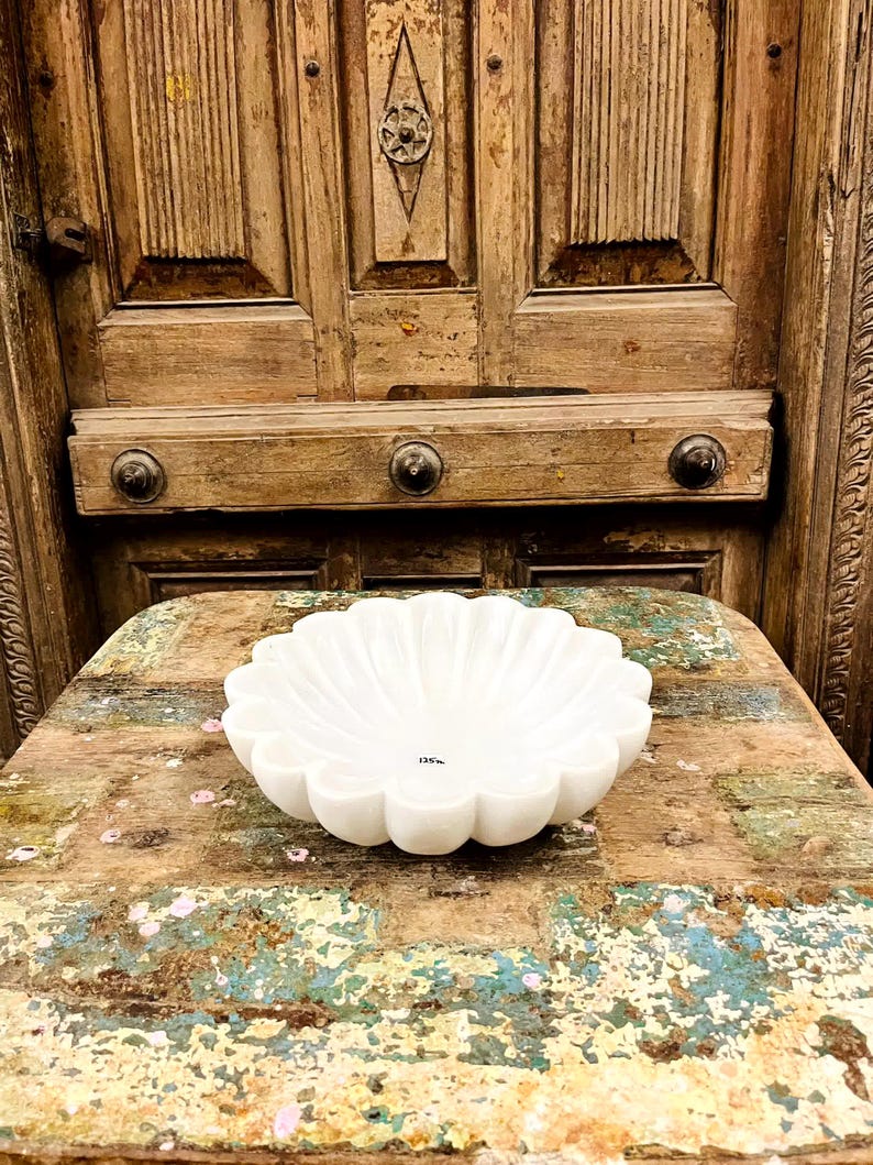 May include: A white marble bowl with a scalloped edge. The bowl has a flower-like shape with rounded edges and a smooth, polished surface. The bowl is on a rustic, multi-coloured wooden surface.