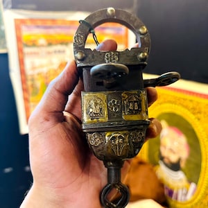 Op de afbeelding: Een antiek slot met ingewikkelde metaalbewerking en een sleutel. Het slot is gemaakt van messing en ijzer en heeft een decoratief ontwerp. Het slot heeft de vorm van een klein doosje met een sleutelgat bovenop. Het slot wordt in een hand gehouden.
