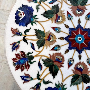May include: A white marble table top with intricate floral inlay. The design features blue, red, green, and brown flowers and leaves with gold-colored stems. The detailed mosaic work creates a decorative surface.