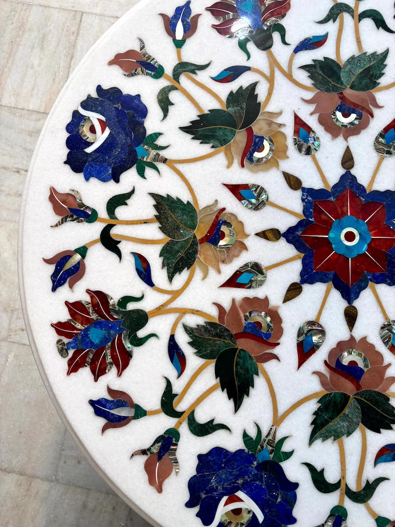 May include: A white marble table top with intricate floral inlay. The design features blue, red, green, and brown flowers and leaves with gold-colored stems. The detailed mosaic work creates a decorative surface.
