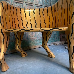 May include: A wooden bench shaped like a tiger, painted gold with black stripes. The bench has four legs and a flat seat.