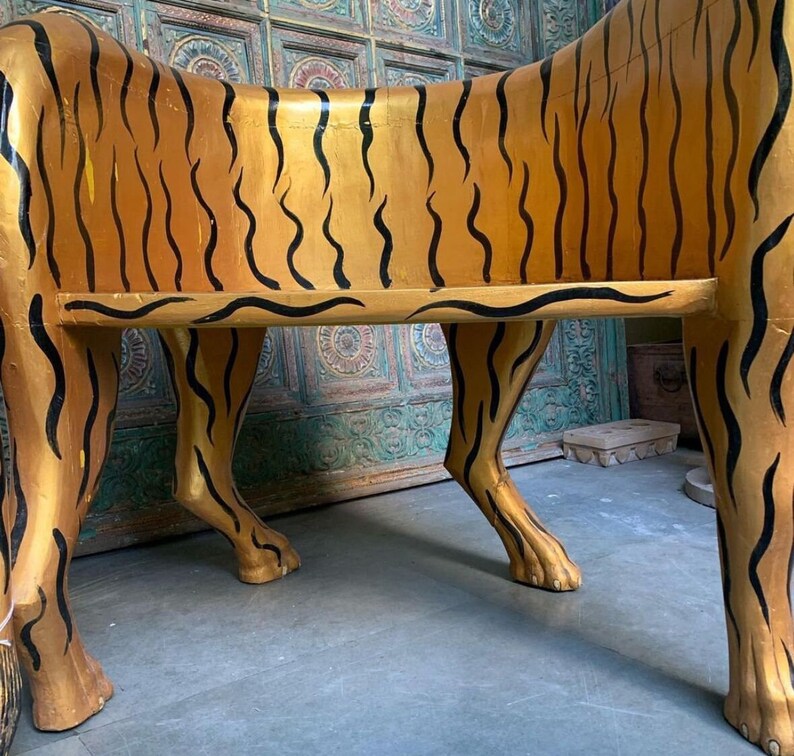 May include: A wooden bench shaped like a tiger, painted gold with black stripes. The bench has four legs and a flat seat.