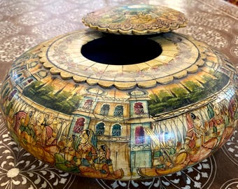 May include: A round, wooden box with a lid, painted with a scene of people and buildings. The box is decorated with intricate carvings and has a dark blue interior.