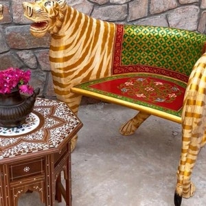 May include: A wooden tiger-shaped chair with a red, green, and gold patterned seat. The chair is hand-painted and has a detailed tiger design. The chair is sitting on a light brown floor next to a small, intricately carved wooden table.