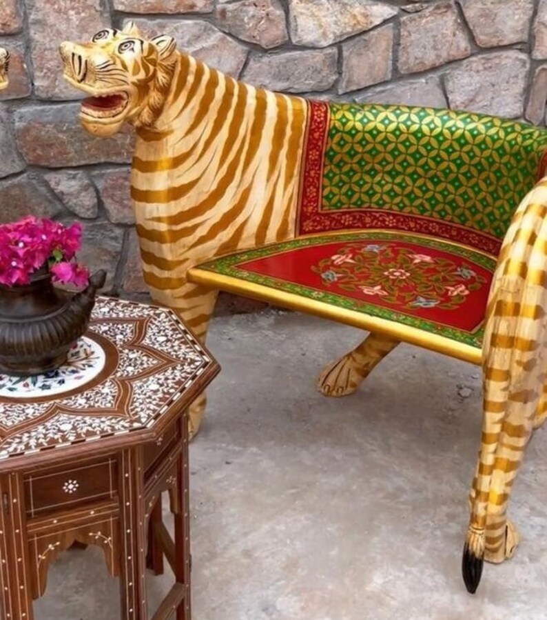 May include: A wooden tiger-shaped chair with a red, green, and gold patterned seat. The chair is hand-painted and has a detailed tiger design. The chair is sitting on a light brown floor next to a small, intricately carved wooden table.